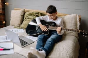 boy sitting on bed with acoustic guitar smiling at a laptop