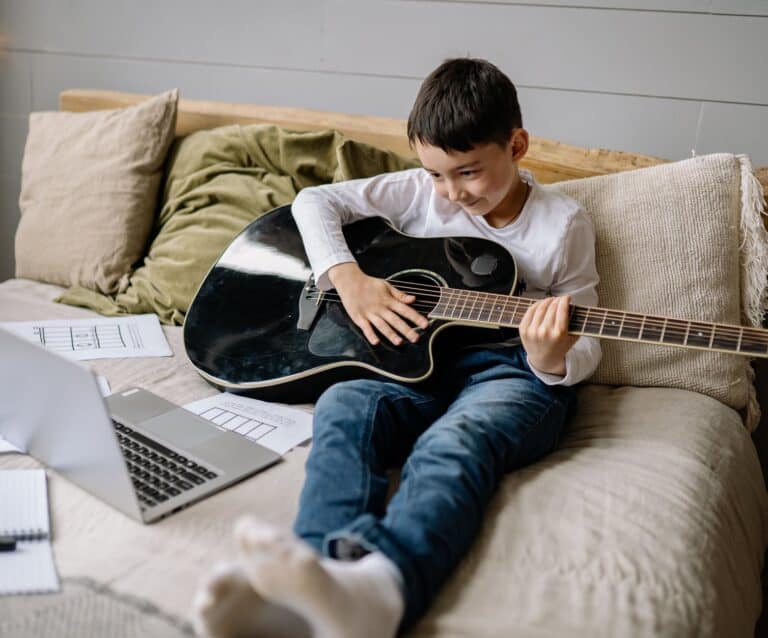 boy sitting on bed with acoustic guitar smiling at a laptop