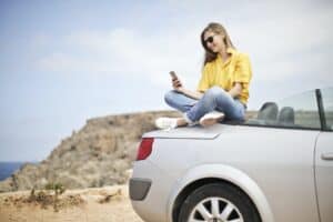 woman in yellow blouse and blue jeans taking selfie while sitting on car