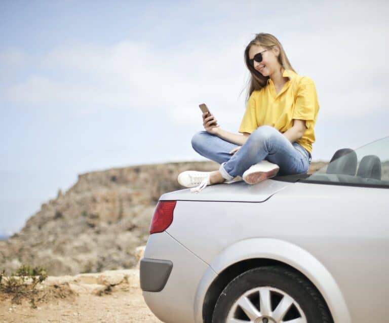 woman in yellow blouse and blue jeans taking selfie while sitting on car