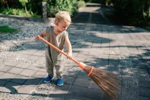 charming child sweeping concrete pavement with broomstick